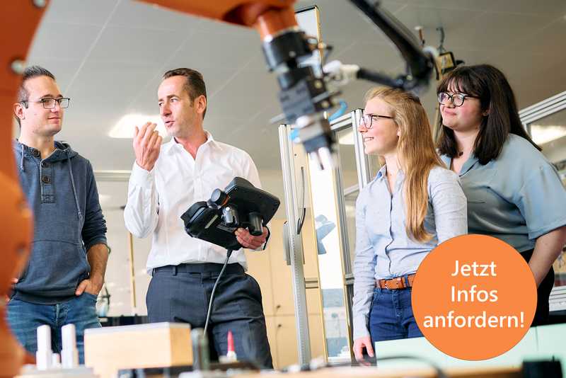 Studierende stehen um einen großen Industrieroboter. Der Professor erklärt etwas und hält dabei ein Steuergerät für den Roboter in der Hand. Der Roboterarm ist im Vordergrund sichtbar und deutet auf eine moderne technische Umgebung hin.