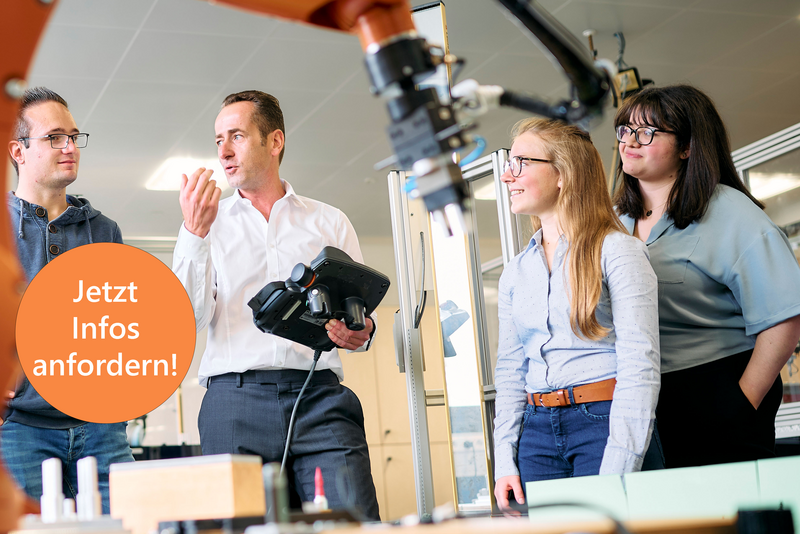Studierende stehen um einen großen Industrieroboter. Der Professor erklärt etwas und hält dabei ein Steuergerät für den Roboter in der Hand. Der Roboterarm ist im Vordergrund sichtbar und deutet auf eine moderne technische Umgebung hin.