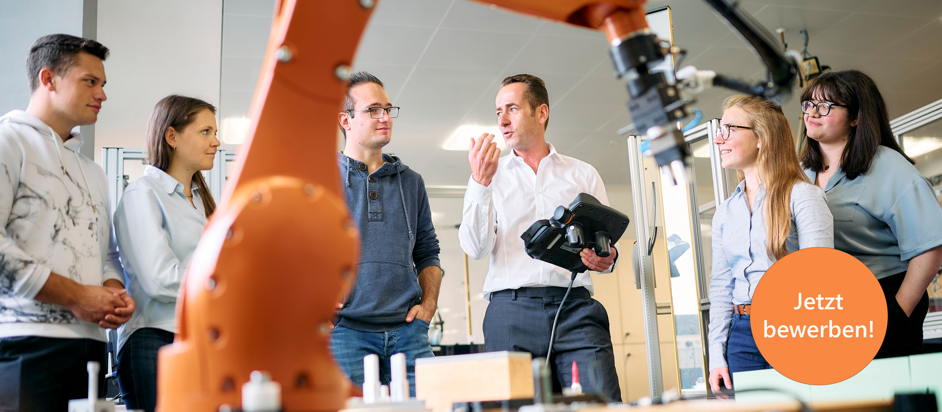 Studierende stehen um einen großen Industrieroboter. Der Professor erklärt etwas und hält dabei ein Steuergerät für den Roboter in der Hand. Der Roboterarm ist im Vordergrund sichtbar und deutet auf eine moderne technische Umgebung hin.