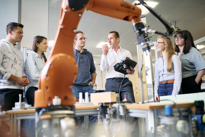 Studierende stehen um einen großen Industrieroboter. Der Professor erklärt etwas und hält dabei ein Steuergerät für den Roboter in der Hand. Der Roboterarm ist im Vordergrund sichtbar und deutet auf eine moderne technische Umgebung hin.