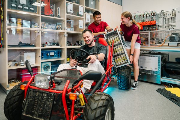 Drei Studierende arbeiten an einem roten Elektro-Kart. Eine Studentin und ein Student heben gemeinsam eine große Batterie, während ein weiterer Mann lächelnd am Steuer des Karts sitzt. Im Hintergrund sind Regale mit elektronischen Bauteilen und technischen Geräten zu sehen.