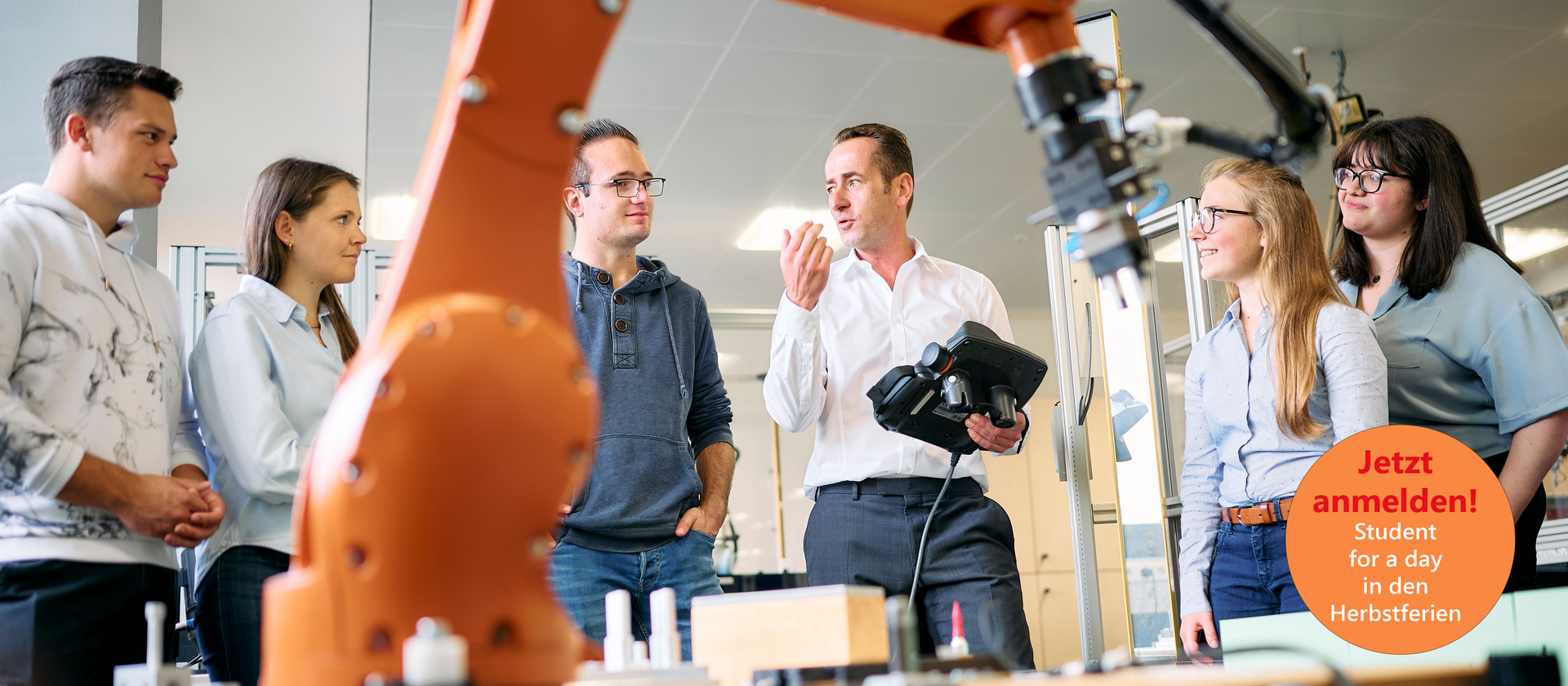 Studierende stehen um einen großen Industrieroboter. Der Professor erklärt etwas und hält dabei ein Steuergerät für den Roboter in der Hand. Der Roboterarm ist im Vordergrund sichtbar und deutet auf eine moderne technische Umgebung hin.