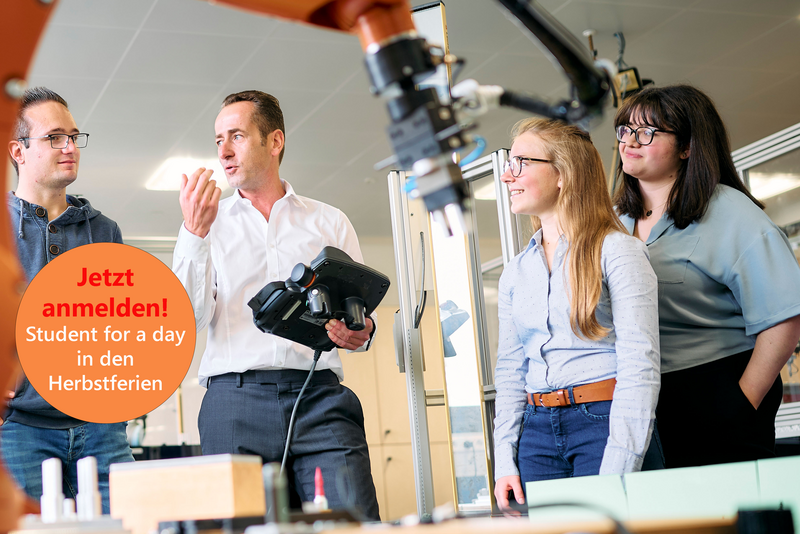Studierende stehen um einen großen Industrieroboter. Der Professor erklärt etwas und hält dabei ein Steuergerät für den Roboter in der Hand. Der Roboterarm ist im Vordergrund sichtbar und deutet auf eine moderne technische Umgebung hin.