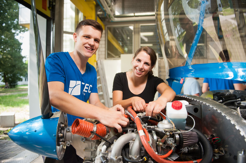 Das Bild zeigt zwei junge Personen, einen Mann und eine Frau, die gemeinsam an einem Motor eines kleinen Flugzeugs arbeiten. Beide lächeln in die Kamera und scheinen mit ihrer Arbeit zufrieden zu sein. Der Mann trägt ein blaues T-Shirt mit der Aufschrift „TEC“, während die Frau ein schwarzes T-Shirt trägt. Sie befinden sich in einer Werkstatt oder einem Hangar, und der Vordergrund zeigt die freiliegenden technischen Komponenten des Flugzeugs. Die Szene vermittelt den Eindruck von Teamarbeit und praxisorientierter technischer Ausbildung, möglicherweise im Rahmen eines Ingenieurprojekts oder einer Lernwerkstatt. Die Umgebung wirkt professionell, aber auch freundlich und kooperativ.