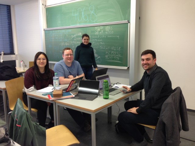 Das Bild zeigt eine Gruppe von vier Personen, die in einem Klassenraum an einem Tisch zusammenarbeiten. Sie haben Laptops vor sich und scheinen sich in einer Arbeitssitzung zu befinden. Im Hintergrund ist eine grüne Tafel zu sehen, auf der einige Notizen oder Berechnungen mit Kreide geschrieben sind. Die Gruppe lächelt in die Kamera, was auf eine entspannte und positive Arbeitsatmosphäre hindeutet. Der Raum wirkt wie ein typischer Hochschulklassenraum, und die Szene vermittelt den Eindruck eines Gruppenprojekts oder einer gemeinsamen Lernsession. Die Studierenden scheinen sich auf eine Aufgabe oder ein Thema zu konzentrieren, das technische oder wissenschaftliche Inhalte umfasst.